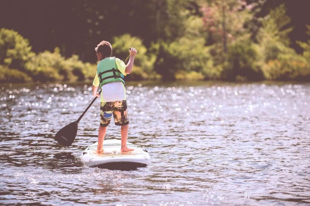 Junge steht auf Stand-up Paddle mit grüner Weste auf dem Wasser von Hinten 