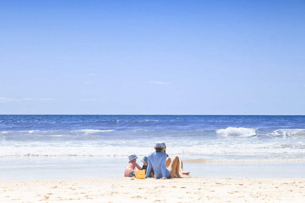 Familienurlaub Tippps am Strand. Eltern mit zwei kleinen Kindern sitzen am Strand vor dem Meer, mit kleinen Wellen im Hintergrund 