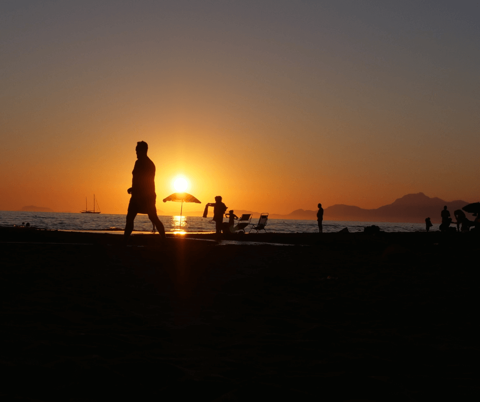 Sonnenuntergang am Strand mit einem Sonnenschirm, verschiedenen Menschen und einem Segelboot am Horizont