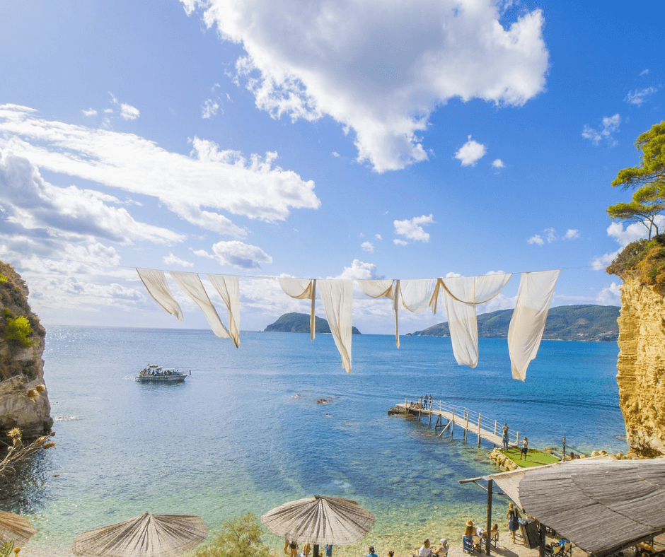 Banana Beach Zakynthos - Strand mit türkisgrünes Wasser, Sonnenschirmen, Steg zum Wasser und vielen Menschen