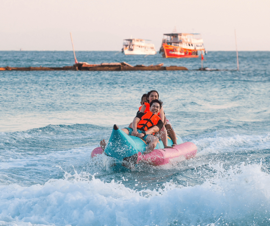 Banana Beach Zakynthos - Drei Personen auf einem Banana Boat mit Schwimmwesten, Schiffe im Hintergrund, Türkisblaues Wasser