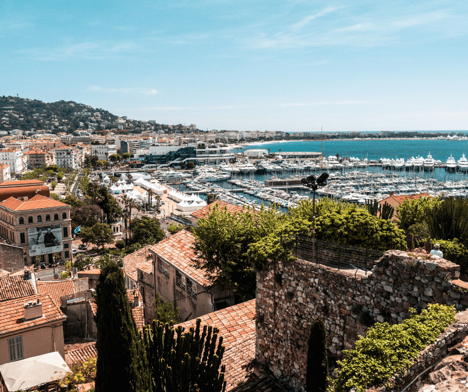 Der Hafen von Cannes mit grün bewachsenen Steinhäusern, weißen Yachten am Steg, einer bergigen Küste und dem blauen Meer im Hintergrund