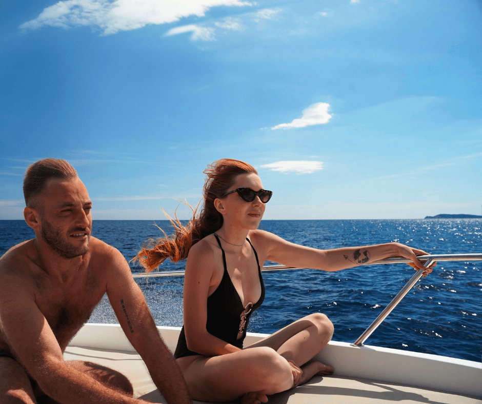Zwei Personen in Badebekleidung an Bord eines Bootes, mit dem Fahrtwind in den Haaren und dem blauen Wasser im Hintergrund 