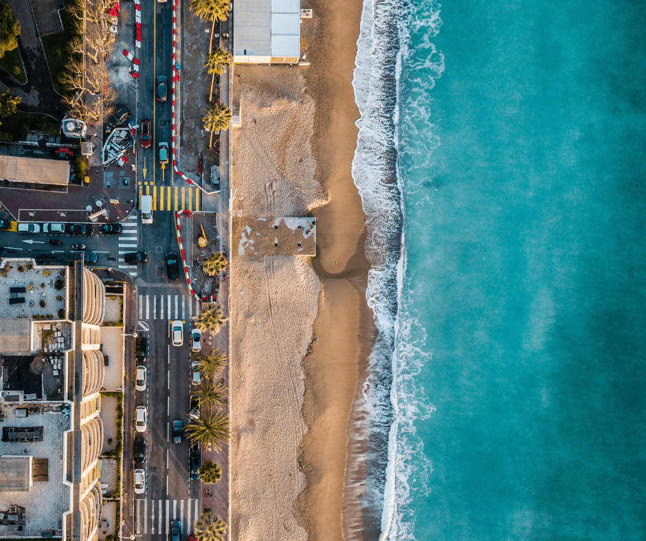 Die Küste von Cannes aus der Vogelperspektive mit dem blauen Meer, dem sandigen Strand und der von Palmen bewachsenen Promenade mit hohen Gebäuden 