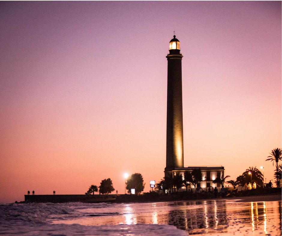 Der Leuchtturm von Maspalomas bei Sonnenuntergang in ein orange-lila Licht getaucht. Im Vordergrund brechen sich die Wellen am Strand. 