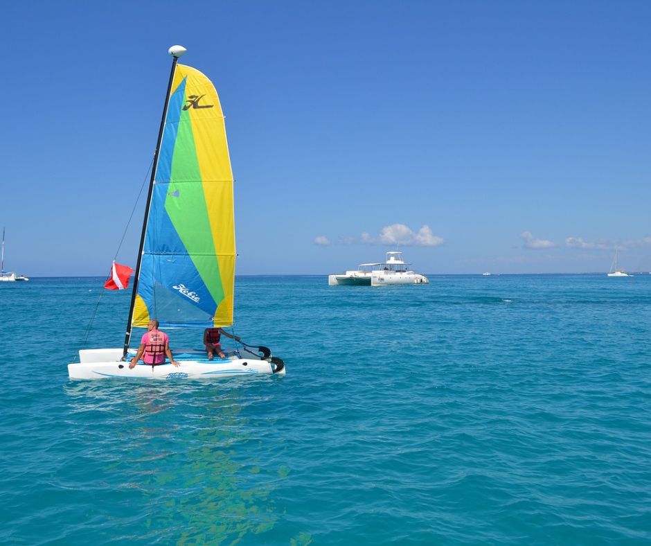 Jolle auf dem Meer. Die Jolle hat ein blau, gelb, grünes Segel und man sieht im Hintergrund weitere Boote.