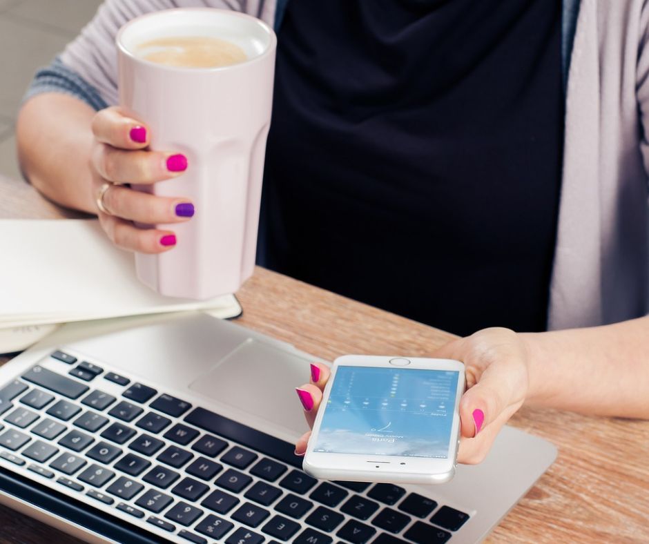 Eine Frau hält in der einen Hand eine Tasse Kaffee und in der anderen ein Smartphone. Vor ihr auf einem Holztisch steht ein Laptop.