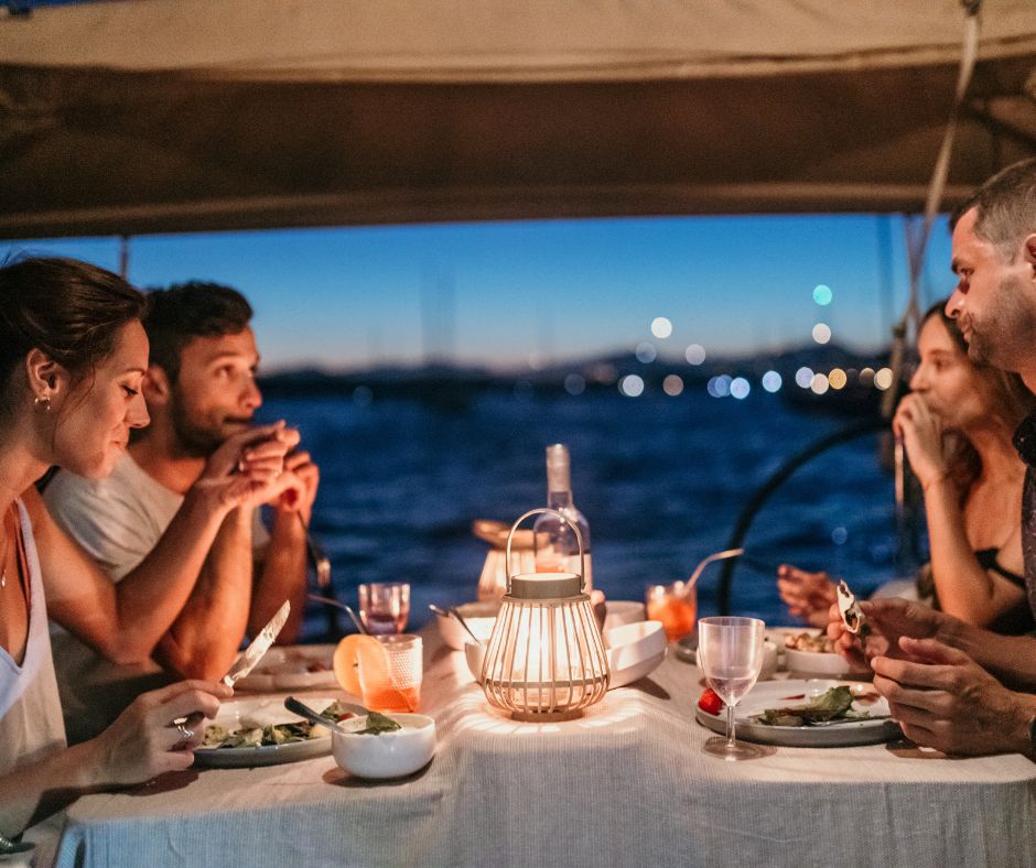 2 Paare sitzen bei Nacht an Bord eines Bootes und genießen ein Dinner bei Kerzenschein.