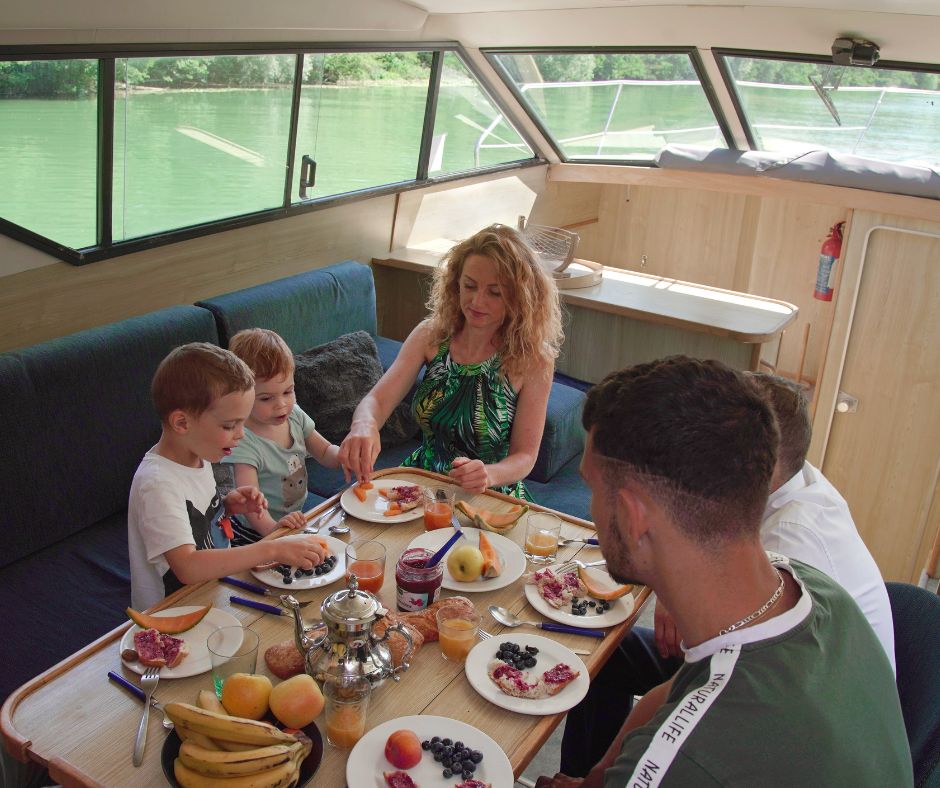 Eine Familie an einem Tisch in einem Hausboot. Links vom Tisch sieht man zwei Männer, der Kamera mit dem Rücken zugewandt. Vor Kopf sitzt eine Frau und links vom Tisch zwei kleine Jungen. Auf dem Tisch befinden sich Teller mit Früchten, Gläser und eine Karaffe – Hausboot günstig mieten.