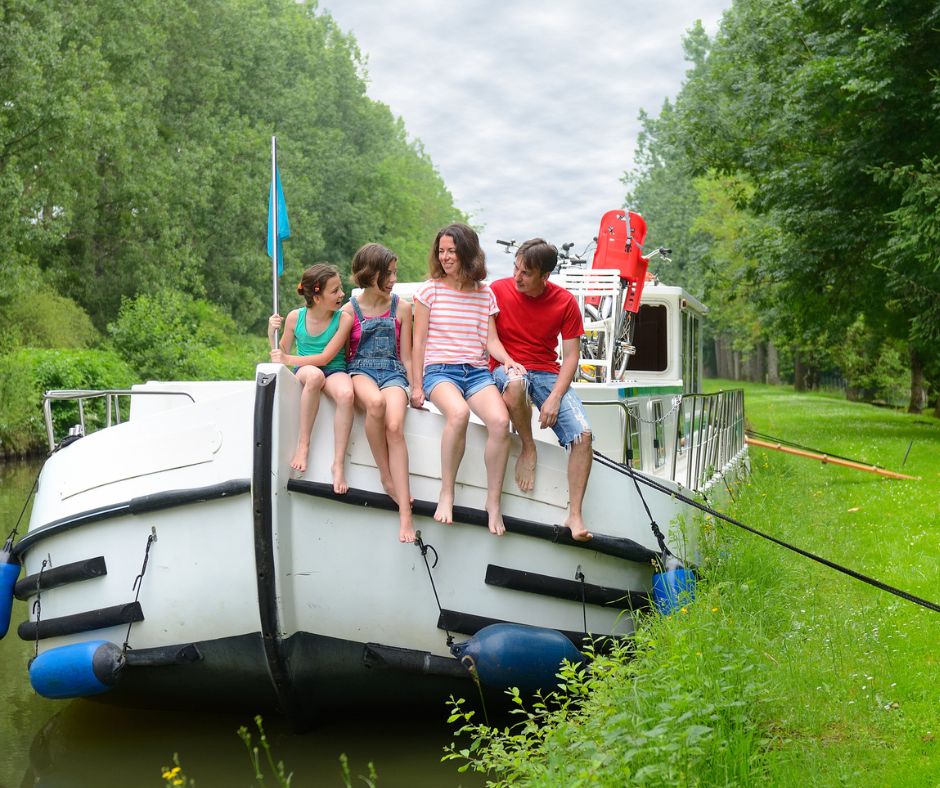 Ein Hausboot, welches auf einem Kanal liegt. Man sieht es von der Vorderseite und eine Familie sitzt am Rand. Zwei Mädchen sitzen links, neben ihnen eine Frau und daneben ein Mann. Im Hintergrund sieht man den Kanal, grüne Wiesen und Bäume.