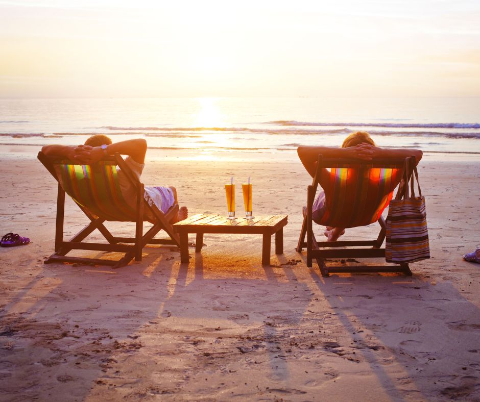 Zwei Personen, die in einem Liegestuhl am Strand sitzen. Zwischen ihnen mittig im Bild sieht man einen Tisch mit zwei Getränken darauf. Im Hintergrund sieht man das Meer bei einem Sonnenuntergang.