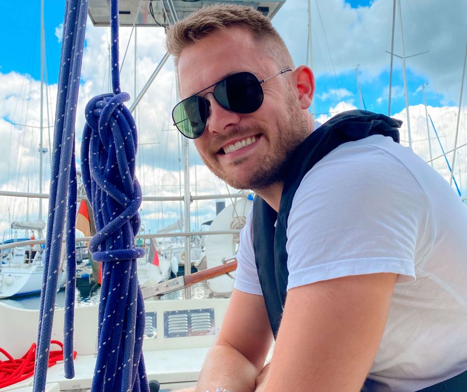 Mann schaut glücklich in die Kamera während er auf einem Segelboot im Hafen sitzt. 