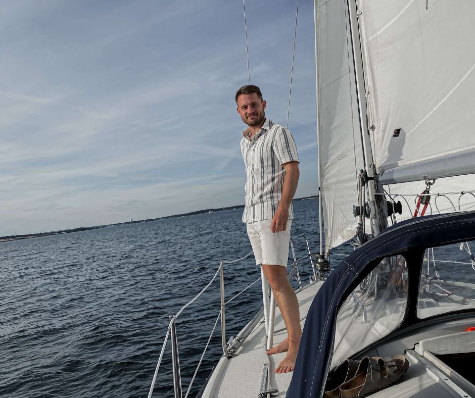 Mann steht an Bord eines weißen Segelbootes. Im Hintergrund das Meer.