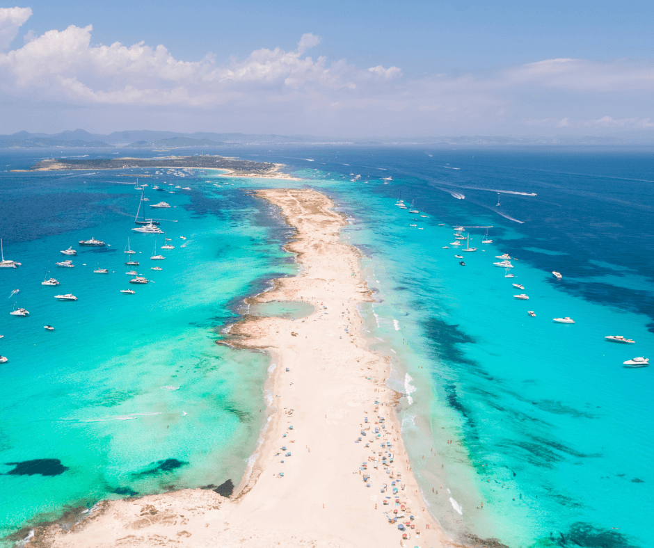 Ein langer, weißer Sandstrand erstreckt sich entlang eines blau-türkisfarbenen Meeres in Ibiza. Zahlreiche Boote sind rund um den Strand zu sehen.