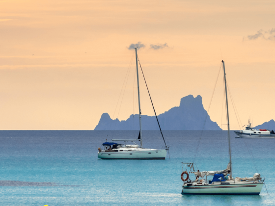Segelboote auf dem Wasser vor Ibiza, während sich ein orangefarbener Sonnenuntergang am Horizont abzeichnet