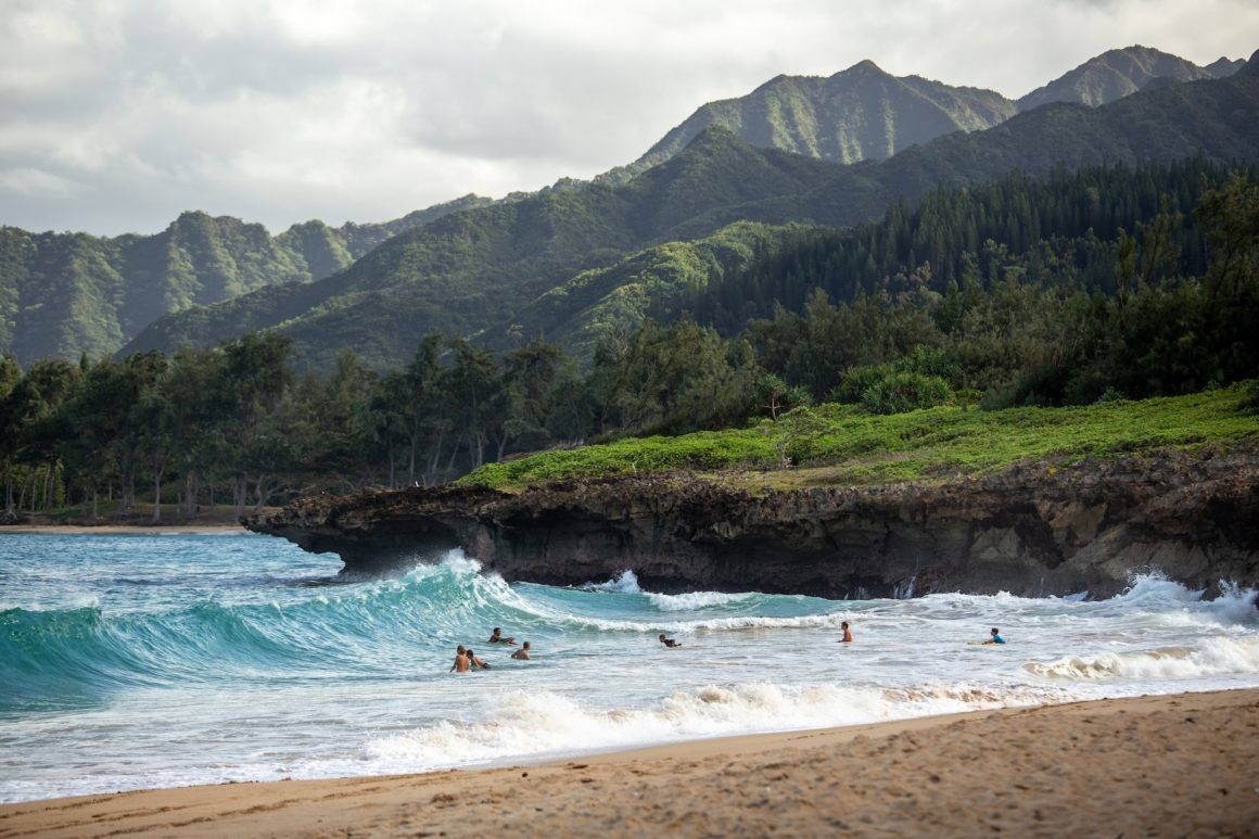 A beach in Oahu, with fine gold sand and people swimming in the water. The ocean waves are crashing into cliffs next to the sea shore, and there are tall green mountains in the distance.