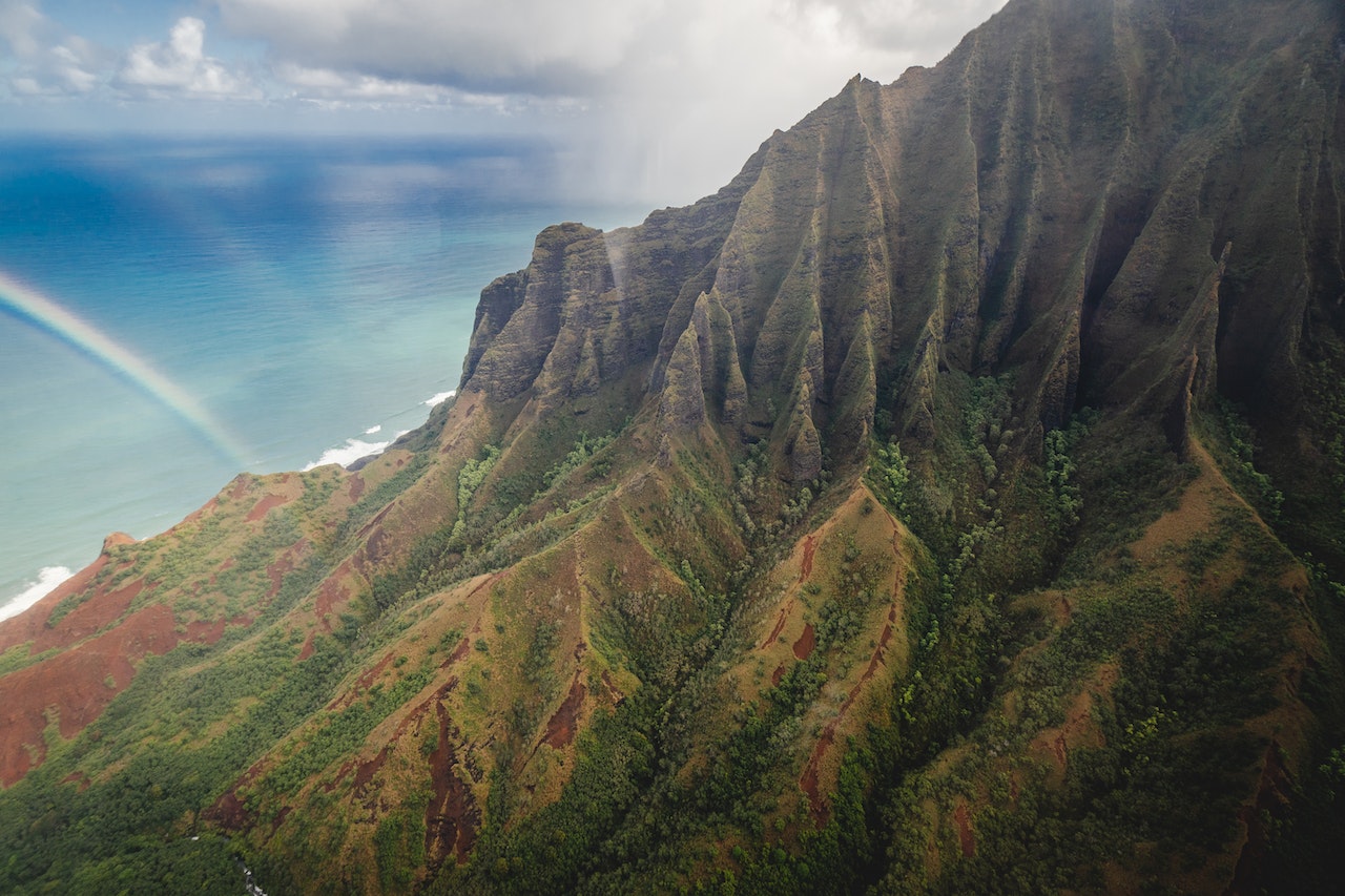 An aerial view of a rainbow arching over the green mountains of Hawaii. with the ocean in the background.