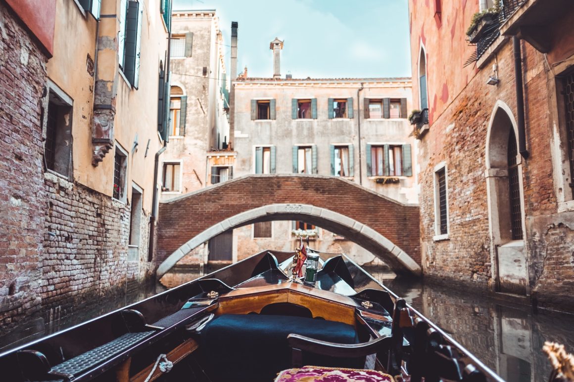 A canal boat in Venice in the forefront that is about to go under an arch bridge, with buildings on either side.