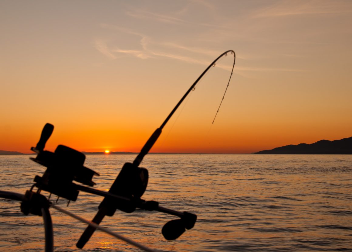 A fishing rod attached to a boat with its reel inside the ocean. The water is calm and the sun is setting.