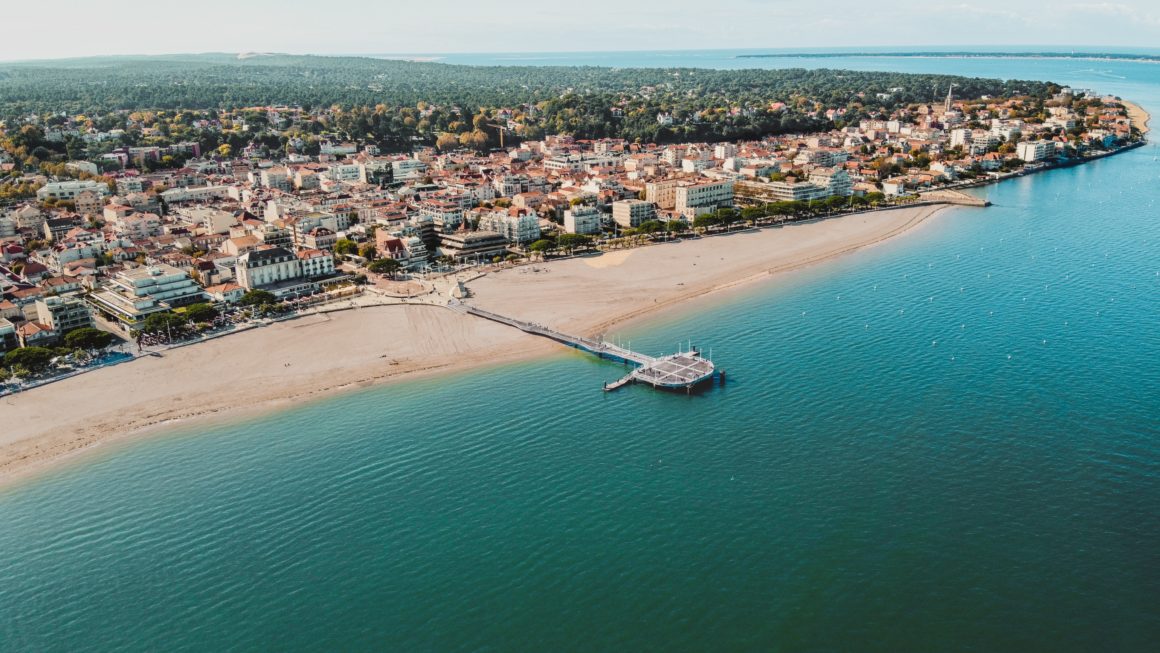 Bird's-eye view of Arcachon. Beach with a pier, with buildings behind. 