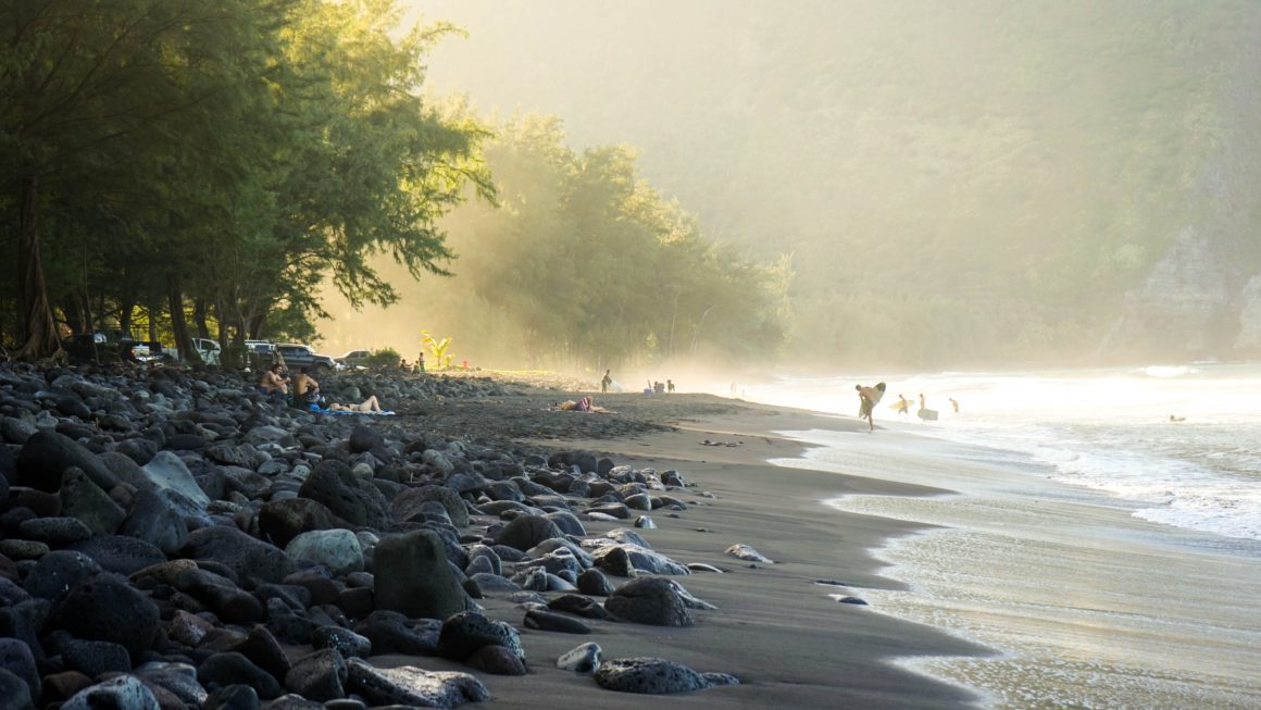 Crashing waves along a black sand beach, with volcanic pebbles and surfers in the back