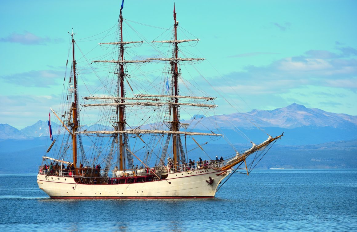 Large white and red Gulet with many people onboard, sailing on blue water with mountain range visible in the background.