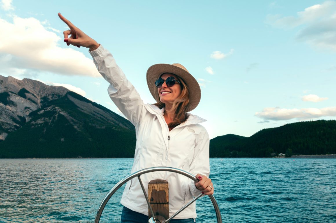 lady at helm of boat sailing and pointing into the distance with blue water and mountain range in the background.