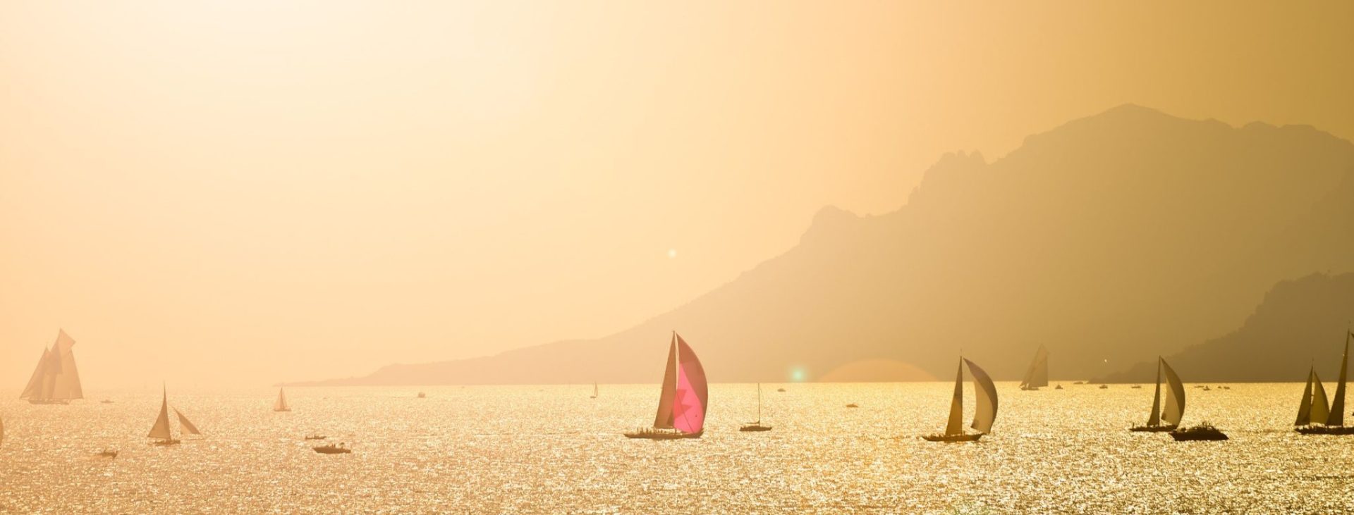 Sailboats and other types of boats sailing in the late afternoon in a bay by some rugged cliffs and mountains