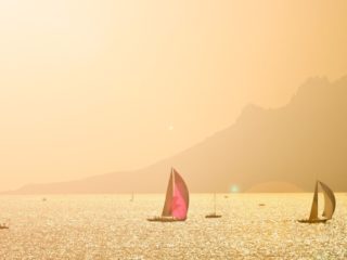 Sailboats and other types of boats sailing in the late afternoon in a bay by some rugged cliffs and mountains
