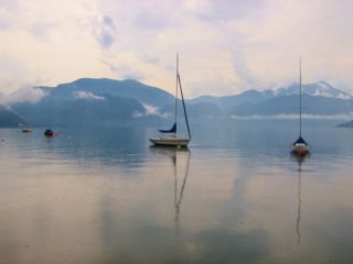 Sailboats moored on a calm lake with misty mountains in the background on an overcast day.