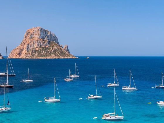 On this picture, there is a rocky landscape in the background with blue turquoise water, where there are sailboats mooring and relaxing