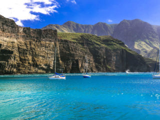 Sailboats anchored on turquoise water beneath towering volcanic cliffs in the Canary Islands on a sunny day.