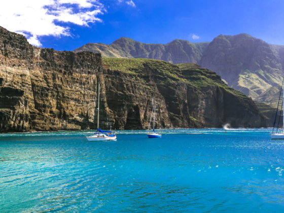 Sailboats anchored on turquoise water beneath towering volcanic cliffs in the Canary Islands on a sunny day.