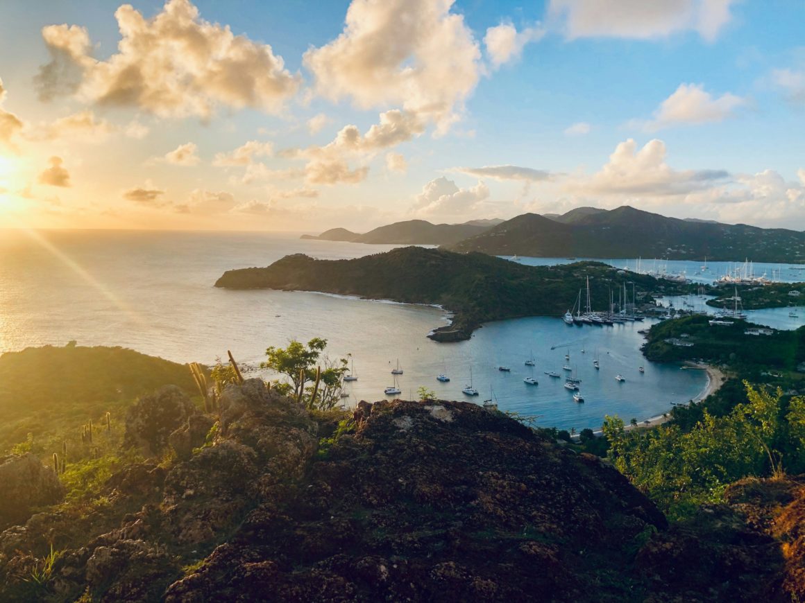 Sunset view over a sheltered bay in the Caribbean, with sailboats anchored and green hills in the background.