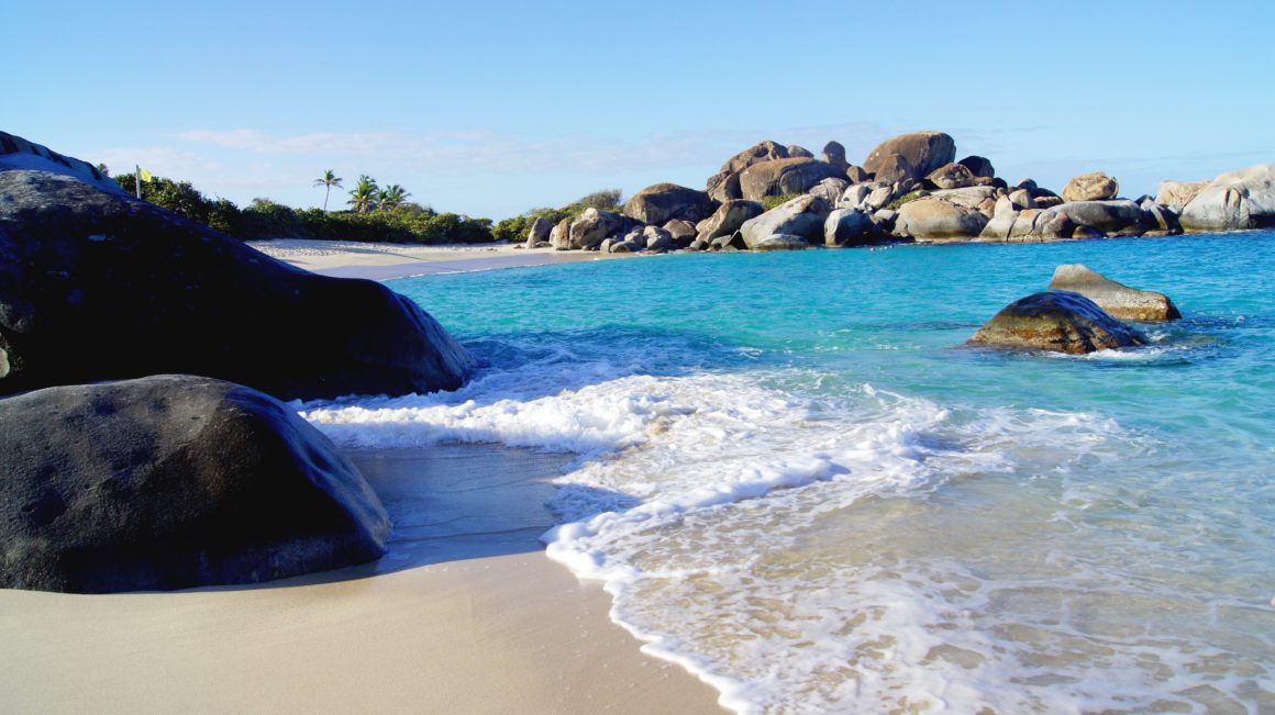Small sandy beach with turquoise waves washing over rocks and large boulders in the Caribbean.