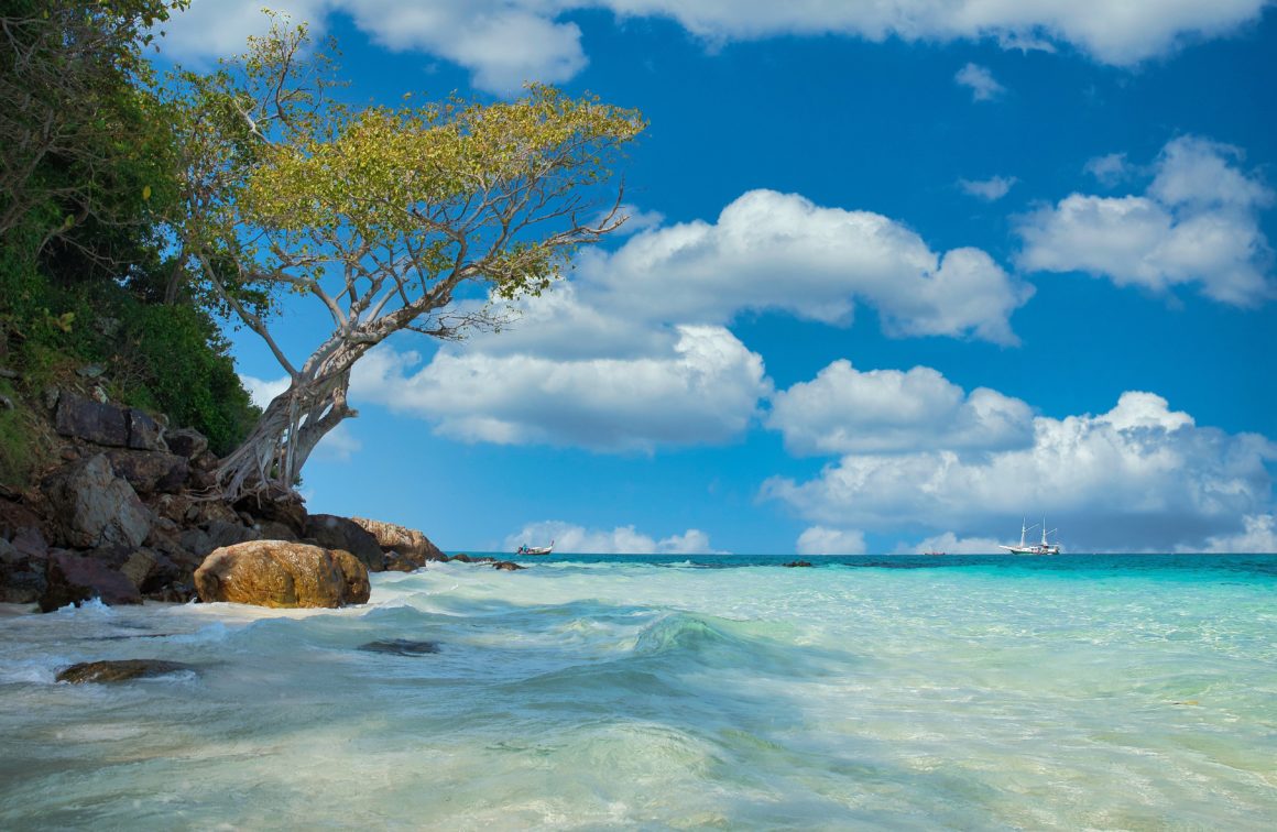 Quiet tropical beach in Thailand with clear water, rocks and a sailboat offshore
