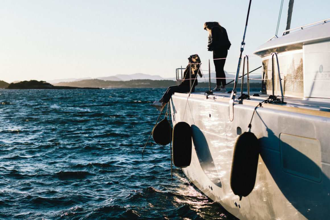 Two people relaxing on the deck of a catamaran sailing on choppy sea at sunset.
