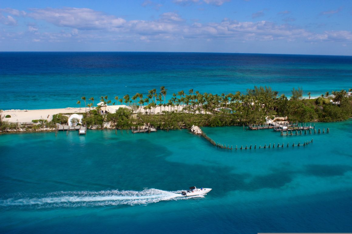 Speedboat cruising through turquoise water past a palm-fringed island and sandy beach in the Bahamas.