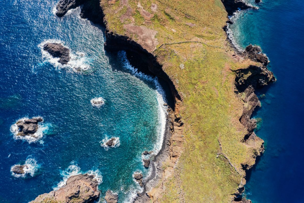 Aerial view of Madeira’s rugged green coastline with dark cliffs and turquoise Atlantic waves crashing on rocky coves.