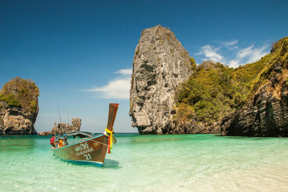 Long-tail boat anchored in shallow turquoise water by steep cliffs in Thailand