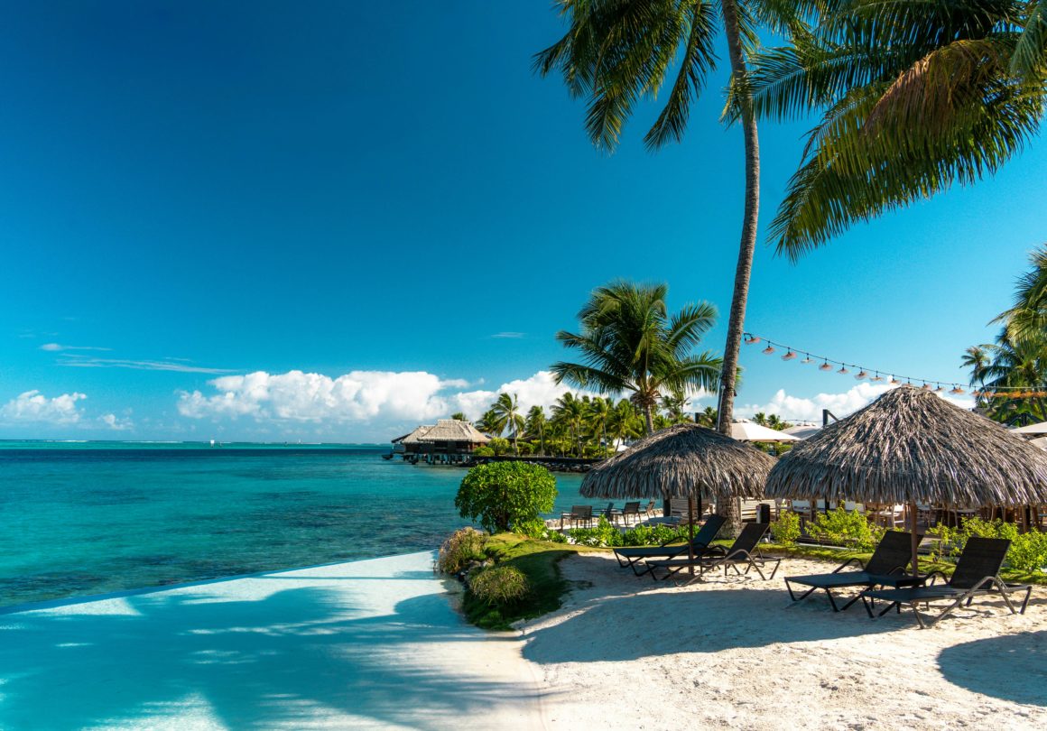 Plage de sable blanc bordée de palmiers avec vue sur des bungalows sur l'eau.