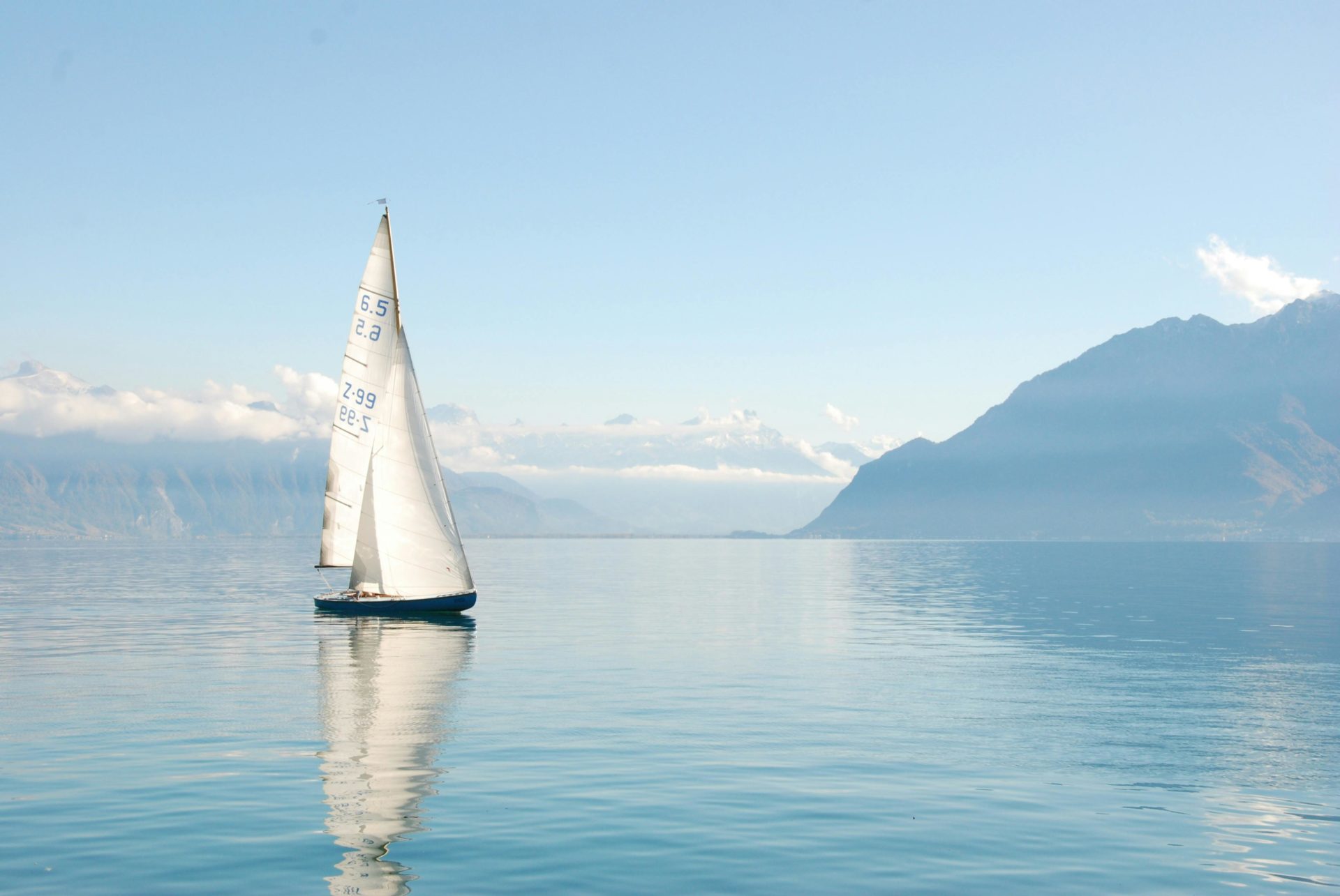 Velero navegando sobre un lago tranquilo con montañas nevadas al fondo.
