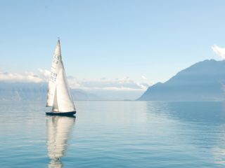 Velero navegando sobre un lago tranquilo con montañas nevadas al fondo.