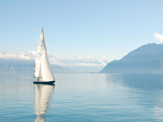 Velero navegando sobre un lago tranquilo con montañas nevadas al fondo.