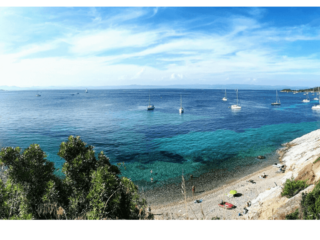 Spiaggia di ciottoli e mare turchese nel sud della Francia, con numerose barche a vela all’ancora al largo.