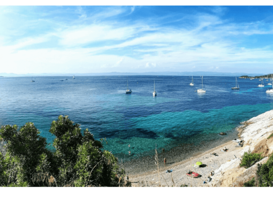 Spiaggia di ciottoli e mare turchese nel sud della Francia, con numerose barche a vela all’ancora al largo.