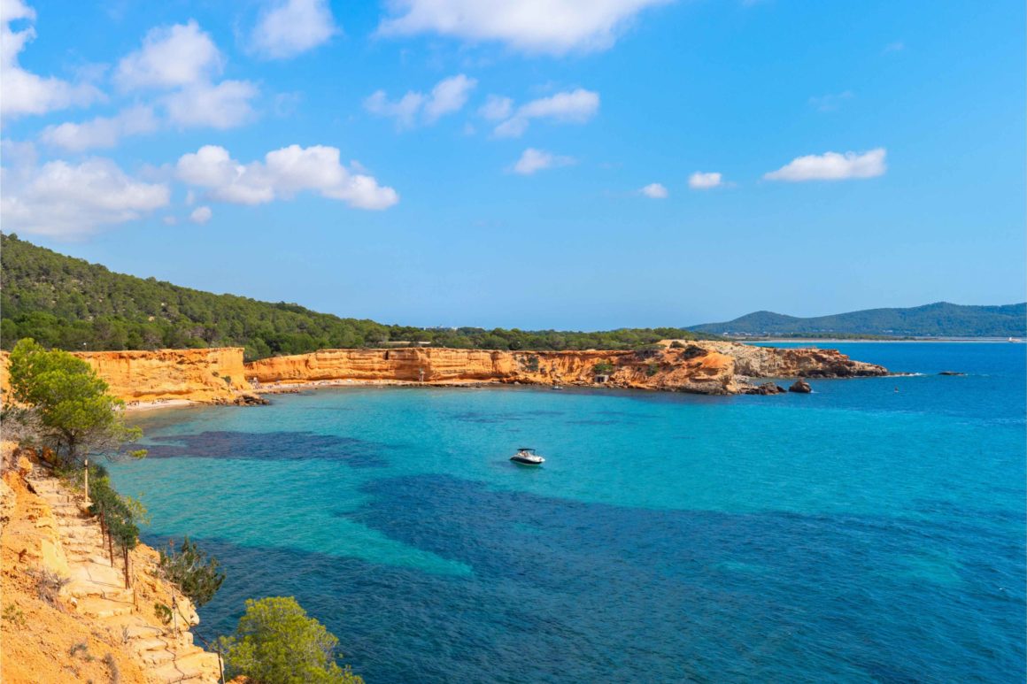 Uitzicht op het strand van Es Bol Nou in het zuiden van Ibiza, warm licht, opaalblauwe zee, een motorboot voor anker in het midden van de baai. Met een van de mooiste ibiza stranden.