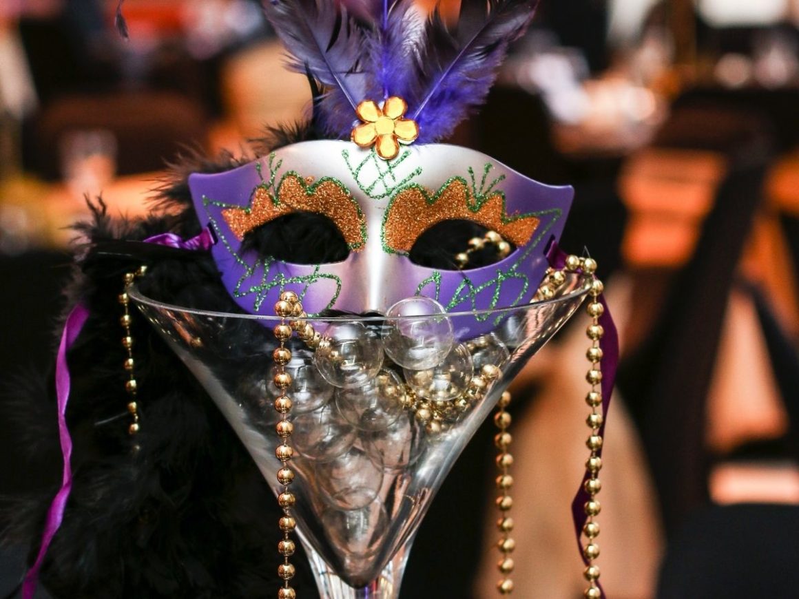 Máscara de carnaval em tons de azul e roxo, dentro de uma taça de vidro. Bolinhas douradas, similares a um colar, poder ser vistas saindo pelas bordas da taça.