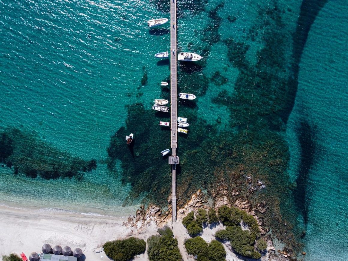 Foto aérea do mar em tons de azul. Uma passarela de madeira liga a praia ao mar e em ambos os lados estão ancorados barcos brancos.