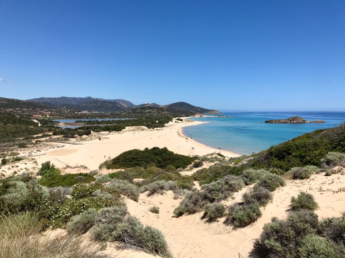 Vista panorâmica de dunas cobertas por areia clara e vegetação nativa. Do lado esquerdo encontra-se o mar em tons de azul e algumas montanhas ao fundo.